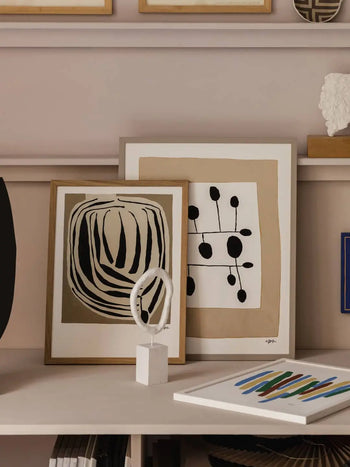 Two framed prints from The Poster Club, including Berit Mogensen Lopezs Improvise (50x70), are displayed on a shelf alongside a small white abstract sculpture and a colorful painting.