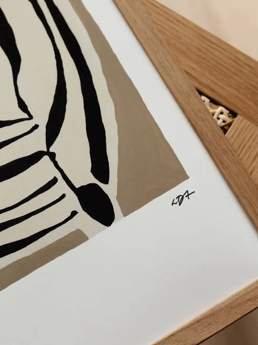 A close-up of The Poster Club Leise Dich Abrahamsen - Layered (50x70) art print features abstract black and beige shapes, framed in light wood, with the artist’s signature in the bottom right corner.