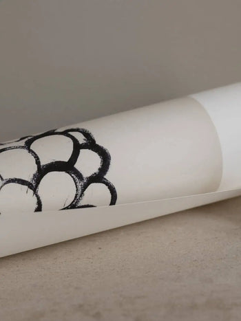A rolled-up sheet of white paper with a black abstract circular pattern, from The Poster Club Madelen Möllard - Flowers in Blue Vase (50x70) art print by The Poster Club, rests on a neutral-colored surface.
