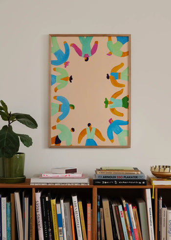 A framed On The Edge (50x70) fine art giclée print by Lise Uhrskov from The Poster Club, showing abstract figures, hangs above a bookshelf with books, magazines, and a small plant on the left end.