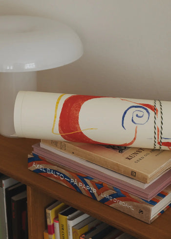 A rolled-up The Poster Club Lucrecia Rey Caro - Nos Encontramos (50x70) art print rests on stacked art books, including Kunst and Axel Salto – På Papir by Lars Dybdahl, atop a wooden shelf beside a white modern lamp.