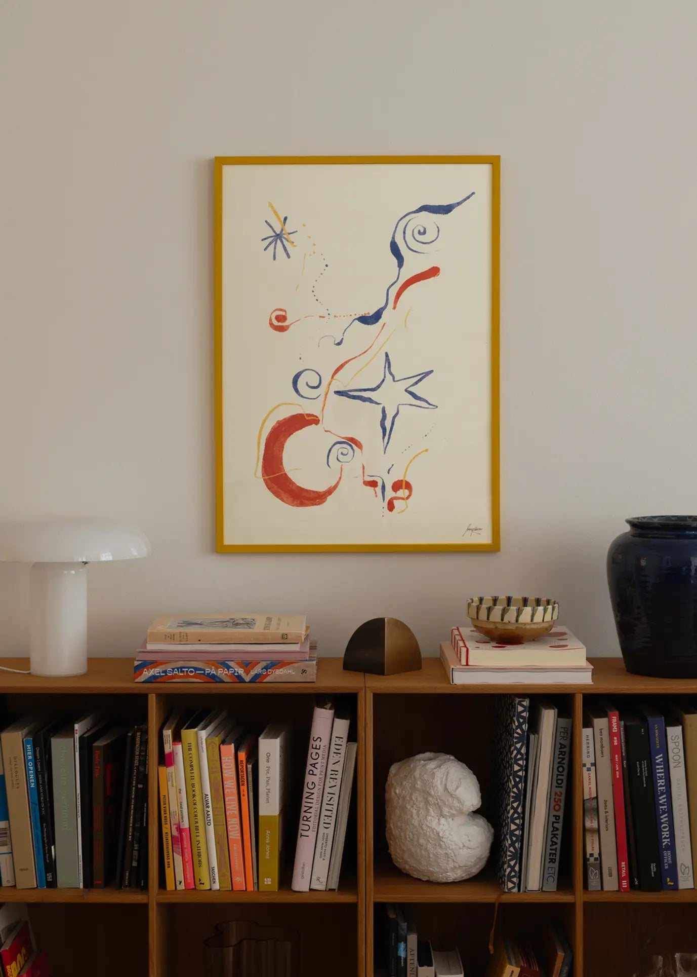 A wooden bookshelf with books and decor sits against a white wall. Above hangs The Poster Club Lucrecia Rey Caro - Nos Encontramos (50x70) art print in a yellow frame, featuring blue and red shapes. A white lamp and dark blue vase top the shelf.