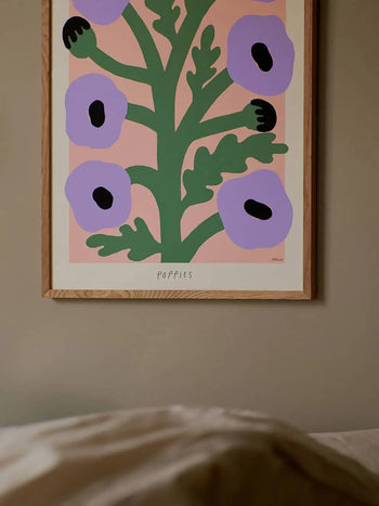The Poster Club Madelen Möllard - Purple Poppies (50x70) is a framed giclée print showing stylized purple poppies with green stems on a pink background, labeled POPPIES at the bottom and displayed above a bed.
