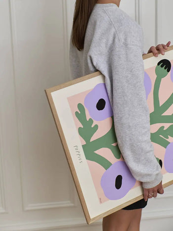 A person in a gray sweater holds The Poster Club Madelen Möllard - Purple Poppies (50x70) giclée art print, showing abstract purple flowers with green stems on a pink background and the word Poppies displayed.