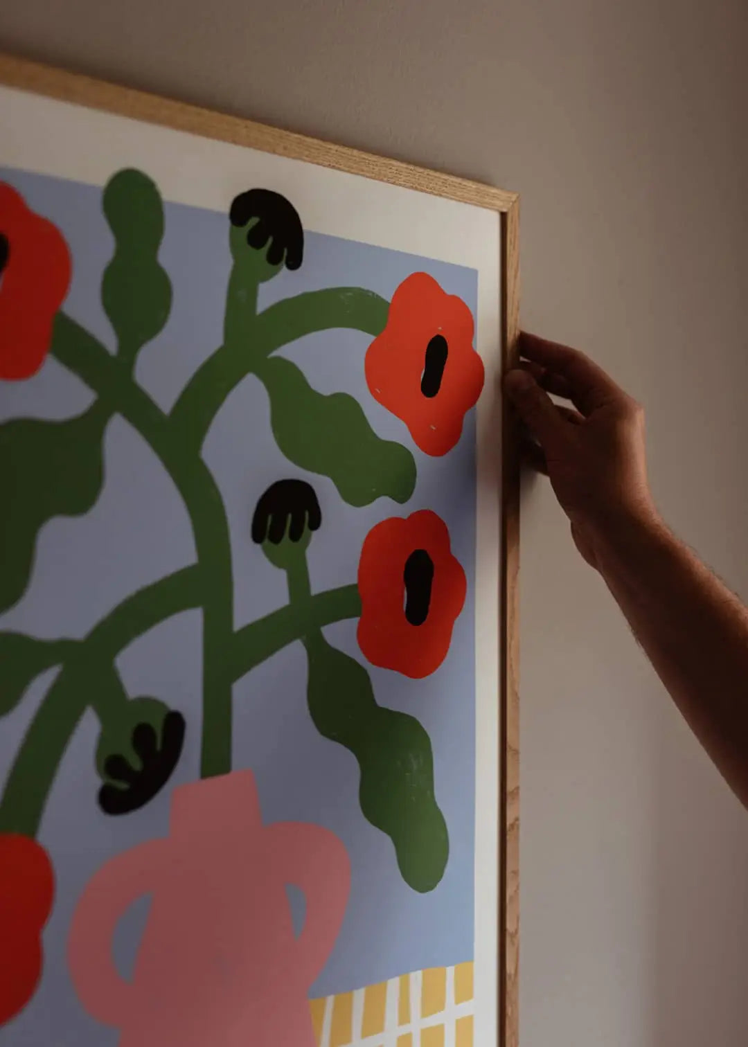 A hand adjusts The Poster Club Madelen Möllard - Red Poppies in Pink Vase (50x70), a bold abstract print featuring red flowers and green stems in a pink vase against a light blue backdrop, on the wall.
