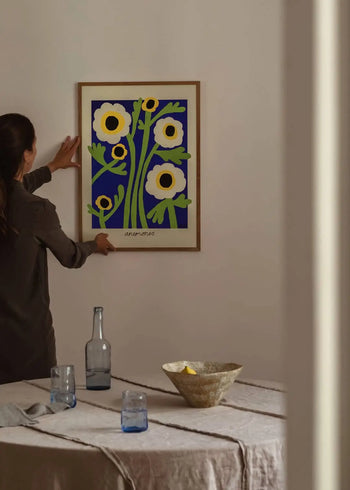 A person hangs The Poster Club Madelen Möllard - Yellow Anemones (50x70) print on a beige wall above a table with a glass bottle, blue glasses, and a woven bowl with a lemon, enhancing the calm, softly lit room.