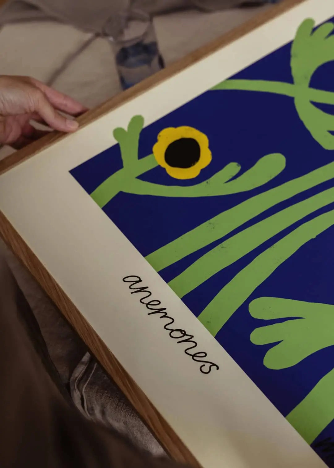 A hand holds The Poster Club Madelen Möllard - Yellow Anemones (50x70) art print, featuring abstract green stems and leaves on a blue background with a single yellow and black flower in a representational style.