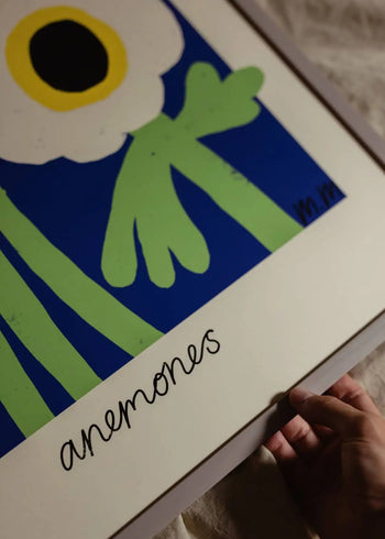 A hand holds The Poster Club Madelen Möllard - Yellow Anemones (50x70) art print, showing a stylized white anemone with a yellow and black center on blue, and anemones written in cursive below the image. Brand: The Poster Club.