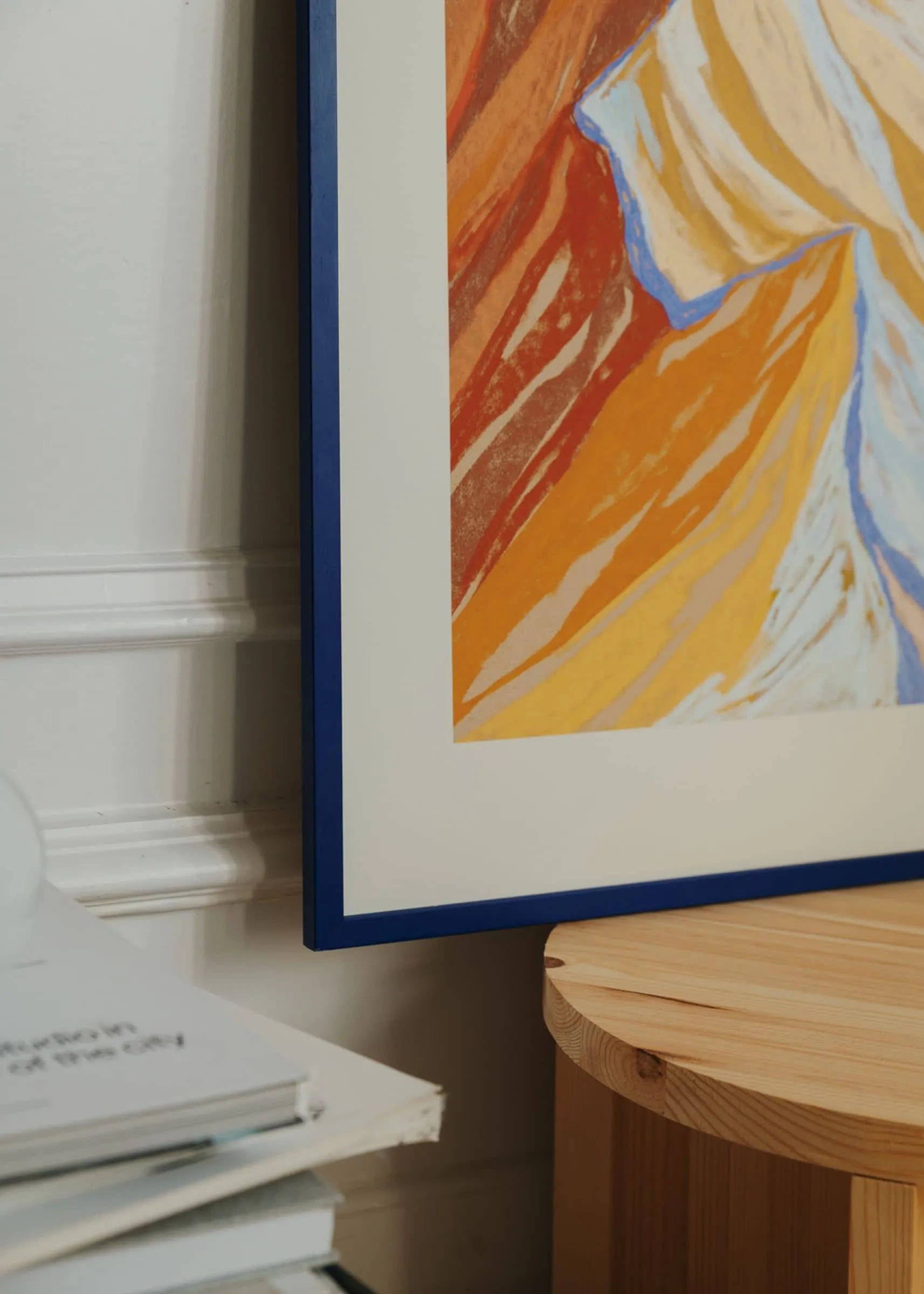 A close-up of The Poster Club’s Mandy Maria - Cuillin Ridge VII (30x40) giclée print in orange, yellow, and blue hangs on a white wall beside a light wooden table and a partially visible book.