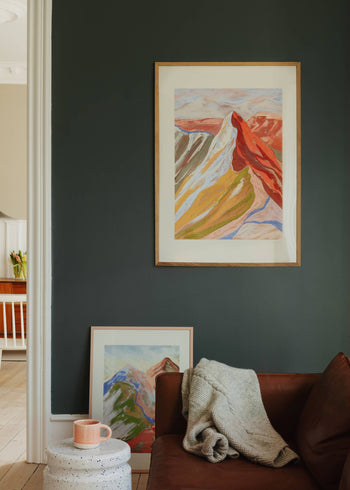 A cozy living room corner features a dark green wall with The Poster Club Mandy Maria - Cuillin Ridge VII (50x70) giclée print framed and hung, another artwork below, a brown sofa with knit blanket, and a small table with a pink mug.