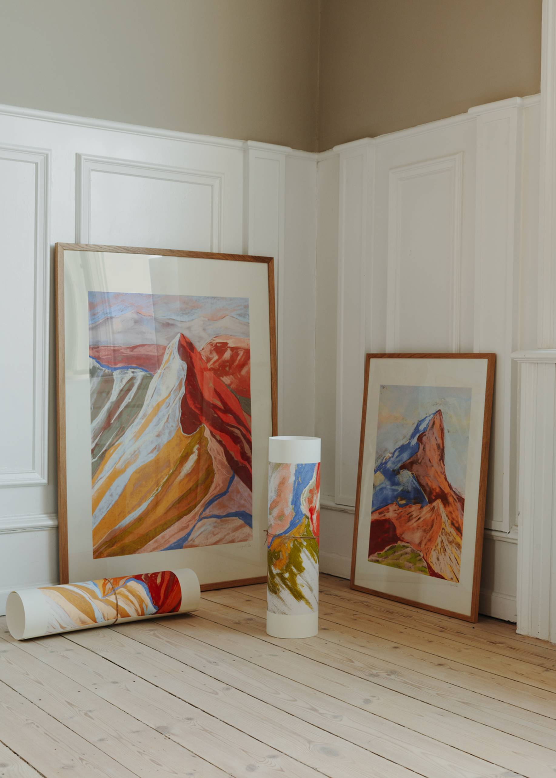 Two framed mountain landscape paintings, including The Poster Club Mandy Maria - Cuillin Ridge VII (50x70), lean against a white paneled wall on a light wooden floor, with two art prints—one rolled, one in a tube—nearby amid soft neutral tones.