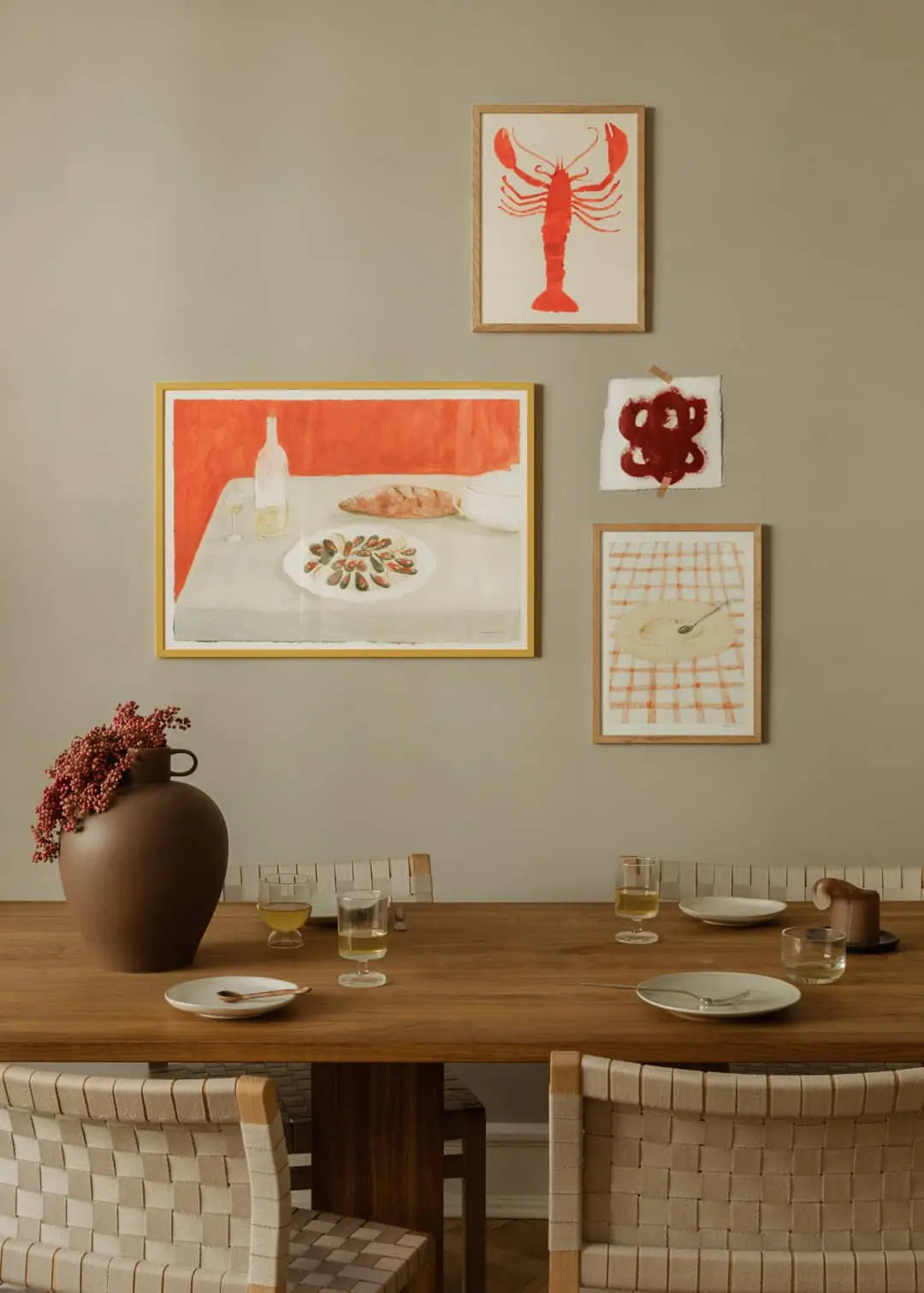 A dining table for four with plates and a brown vase of pink flowers sits beneath framed art prints, including Bloom by Berit Mogensen Lopez from The Poster Club, alongside prints of a red lobster, fish, and an abstract table scene.
