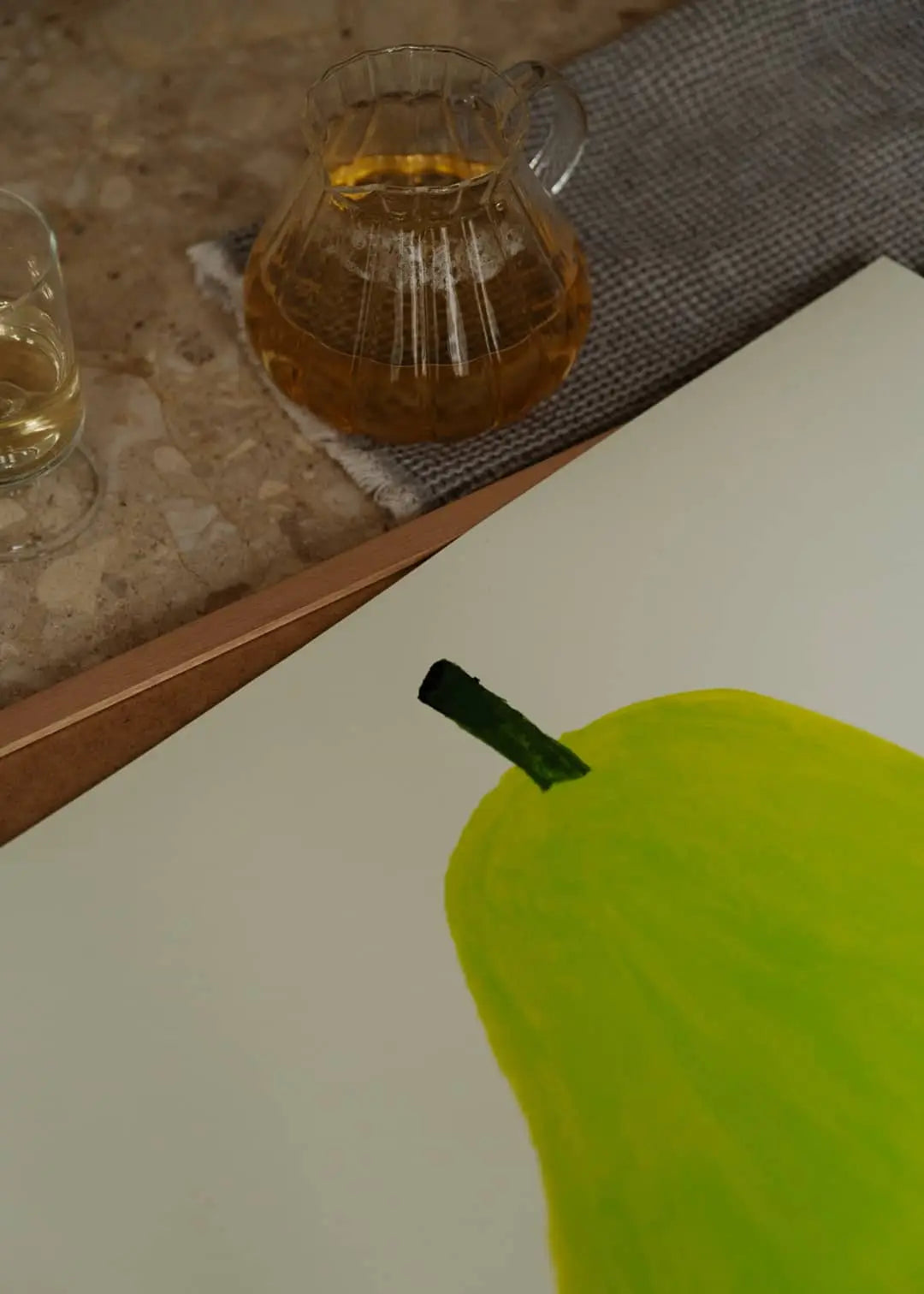 A glass and a carafe with a light yellow drink sit beside The Poster Clubs Marina García Fernández - Pera Limonera 1 (50x70) art print, which features the upper half of a green pear on white paper.