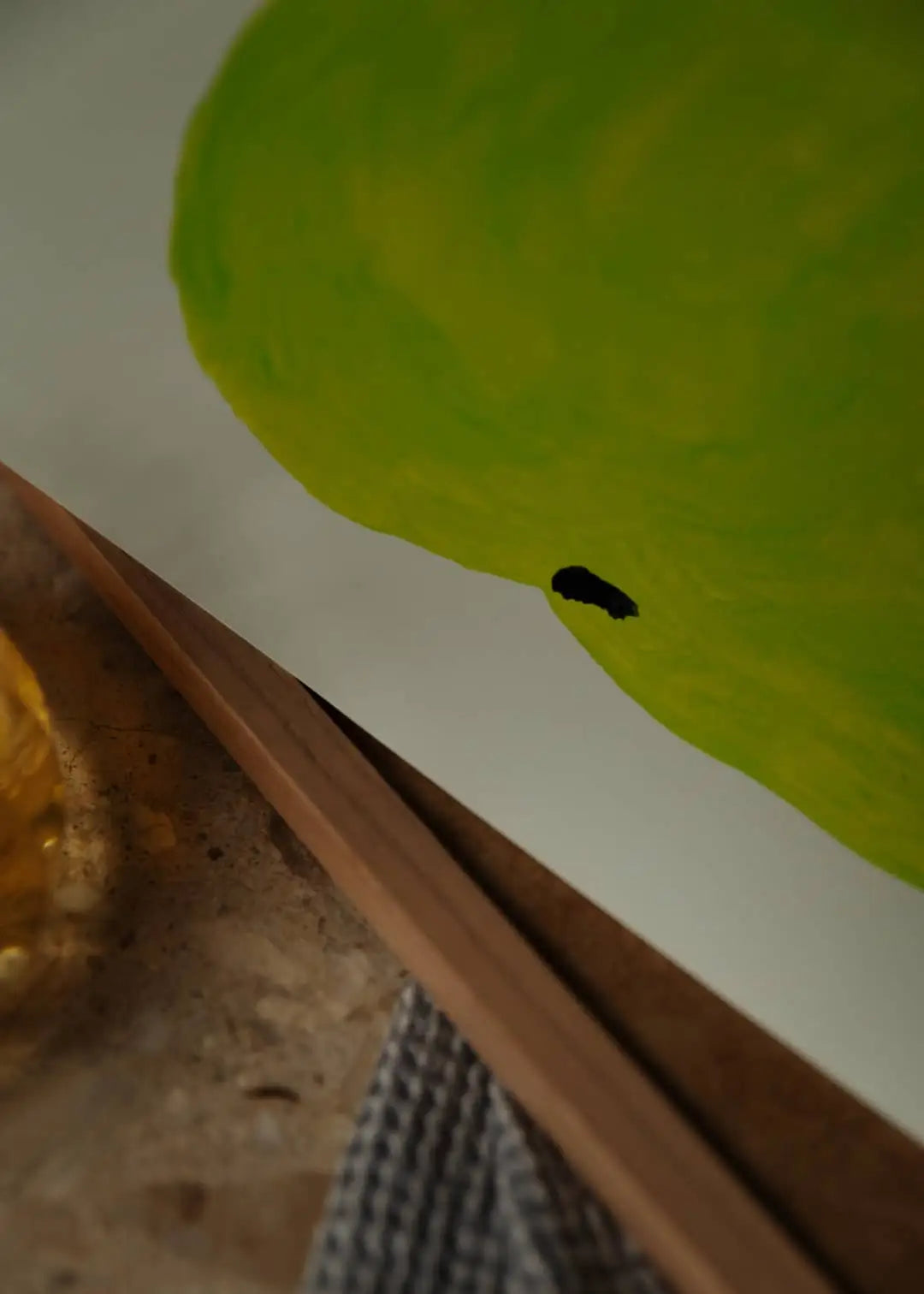 A close-up of The Poster Club’s Marina García Fernández - Pera Limonera 1 (50x70) art print, showing a large green shape with a black spot on white paper, partly on a wooden frame and textured surface.