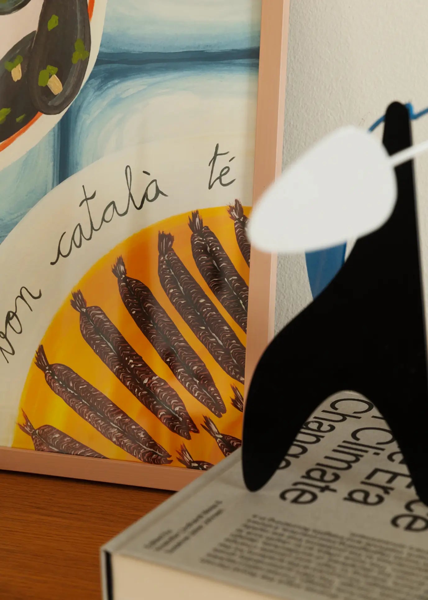 A framed The Poster Club Marta Leyva - Bona Cuina 01 (30x40) print leans against the wall, complemented by a black and white abstract sculpture and The Climate book on a wooden surface for a Mediterranean vibe.