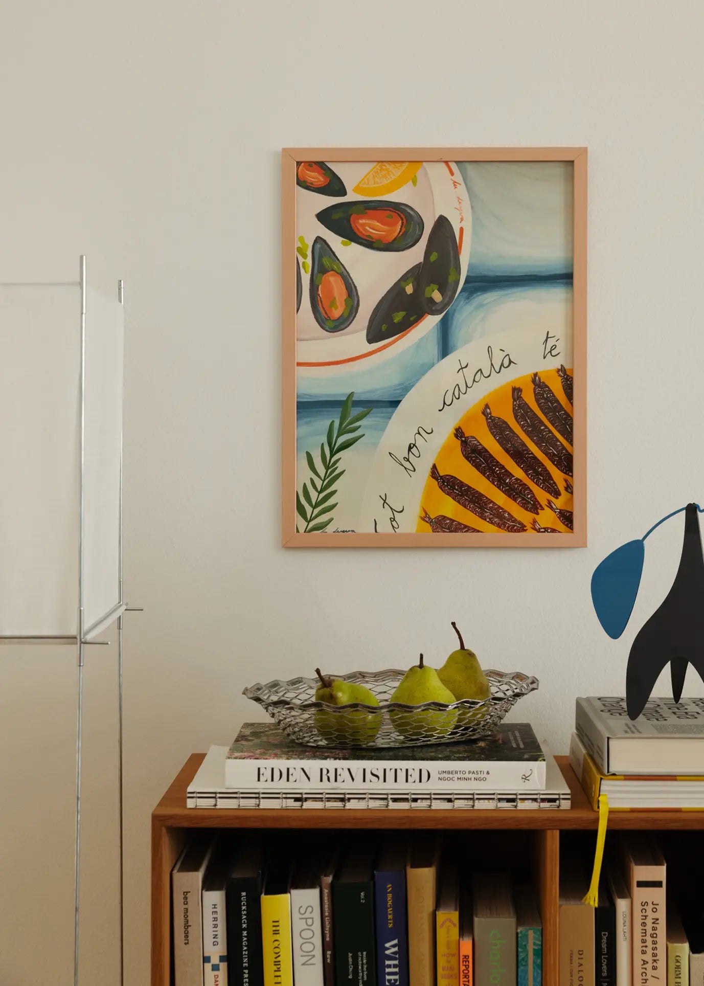 A cozy scene features The Poster Clubs Marta Leyva - Bona Cuina 01 (30x40) giclée print above a bookshelf with pears, books, and a mobile. Muted tones and natural light create a relaxed Mediterranean feel.