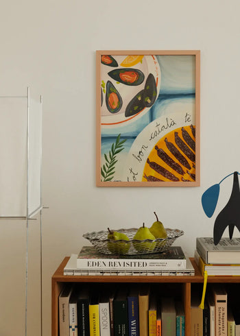 A cozy scene features The Poster Clubs Marta Leyva - Bona Cuina 01 (30x40) giclée print above a bookshelf with pears, books, and a mobile. Muted tones and natural light create a relaxed Mediterranean feel.