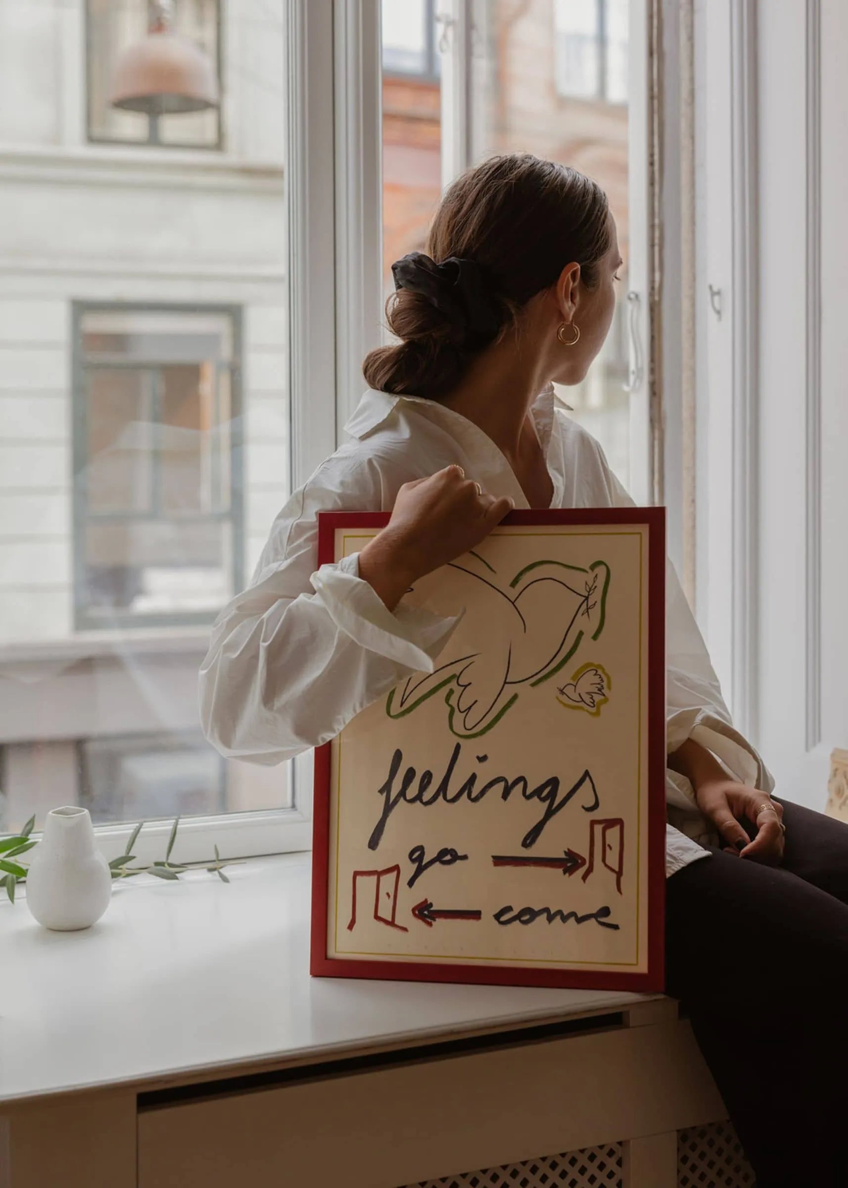 A woman sits by a window, holding The Poster Club’s Marta Leyva - Feelings (30x40) Giclée print featuring a dove, “feelings go come” text, and directional arrows. A white vase with green leaves on the sill adds to the Mediterranean vibe.