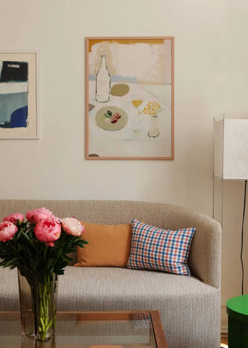 A beige sofa with an orange cushion and plaid pillow sits beneath The Poster Clubs Checkered art print by Berit Mogensen Lopez. Pink flowers in a glass vase rest on a glass coffee table, with a tall white lamp against the cream wall.