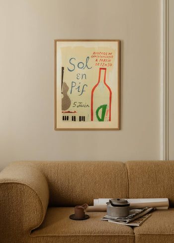 A beige curved sofa sits by a light wall with The Poster Club Max Ososki - Sol En Pif (30x40) art print above, showing a collage of guitar, bottle, and piano keys with French text. A tray on the sofa holds a teapot, cup, and magazines.
