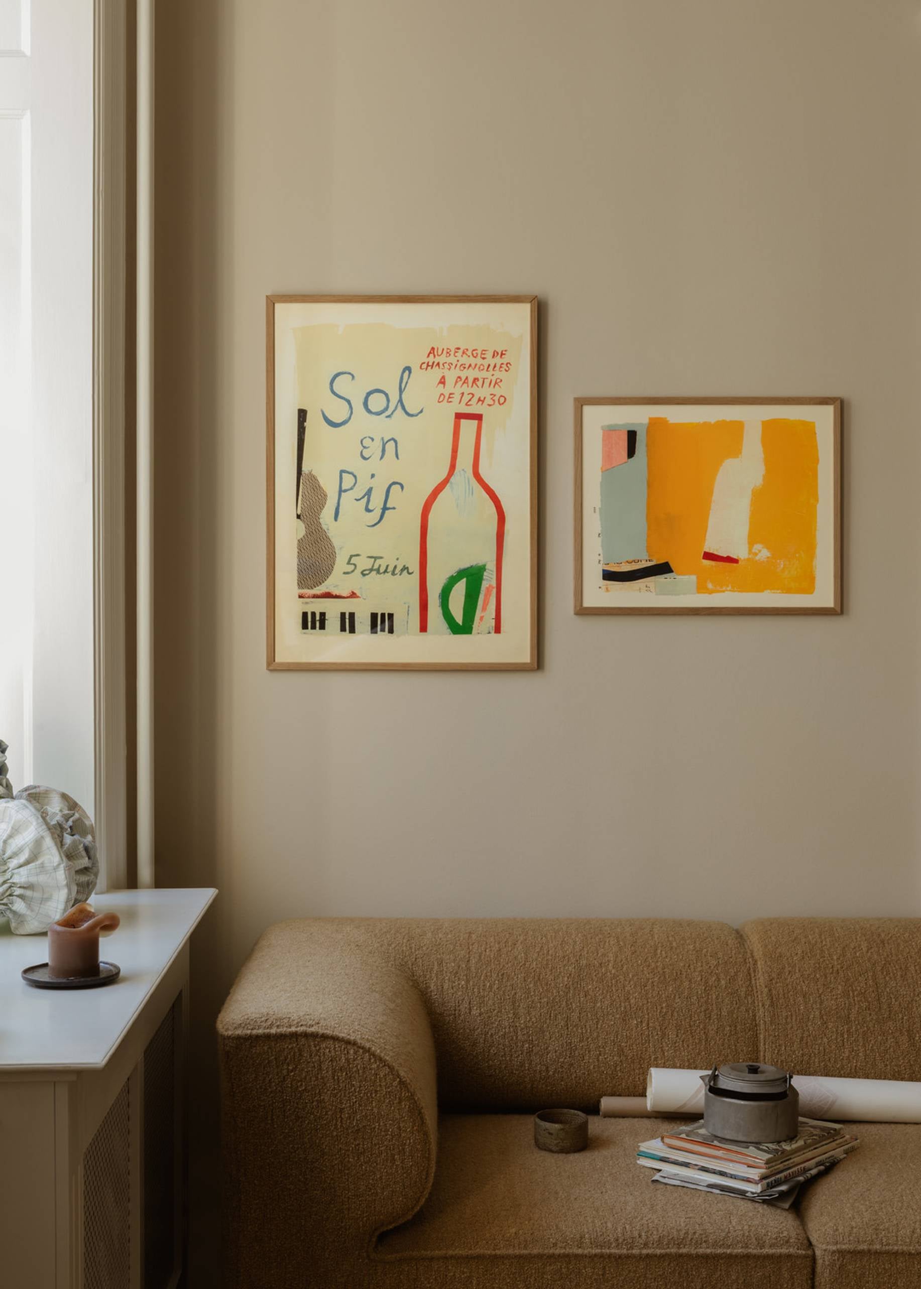 A beige sofa sits below two framed Sol En Pif art prints by Max Ososki from The Poster Club on a tan wall; nearby, a white windowsill displays a glass vase and fan-shaped decor. Magazines and art supplies are stacked on the sofa.