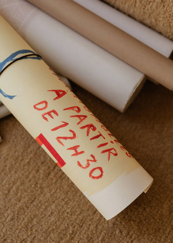 A rolled-up Poster Club Max Ososki - Sol En Pif (30x40) print rests on a brown carpet beside other cylindrical rolls, featuring French red handwritten text reminiscent of Ososki’s collage style.