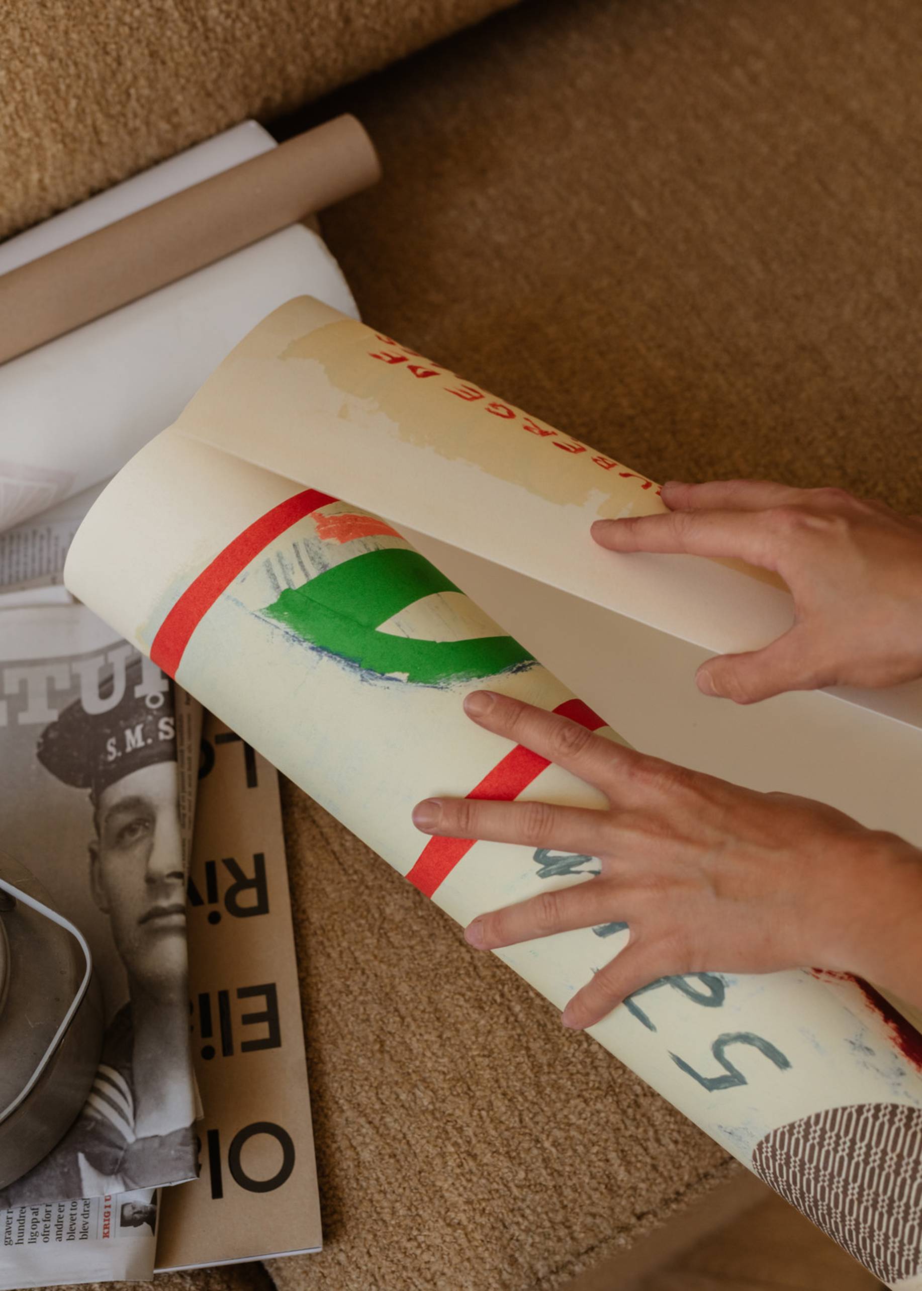 Someone unrolls vibrant art prints on a brown surface beside cylindrical cardboard tubes and a black-and-white magazine featuring The Poster Club Max Ososki - Sol En Pif (30x40) by The Poster Club.