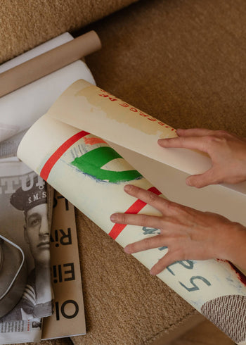 Someone unrolls vibrant art prints on a brown surface beside cylindrical cardboard tubes and a black-and-white magazine featuring The Poster Club Max Ososki - Sol En Pif (30x40) by The Poster Club.