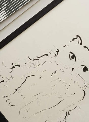 A minimalist black ink drawing of a fluffy wide-eyed cat on white paper with a black border, The Poster Club Oamul - Cat (50x70) Poster is seen at an angle with part of another striped object in the corner—a unique art print by The Poster Club.