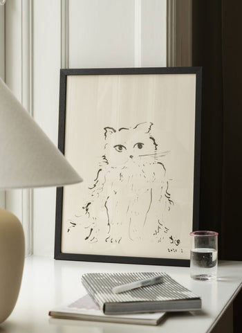 The Poster Club Oamul - Cat (50x70) poster by The Poster Club, framed in black, is displayed on a white table with a notepad, pen, glass of water, and beige lamp. The setting has white and dark brown walls in the background.