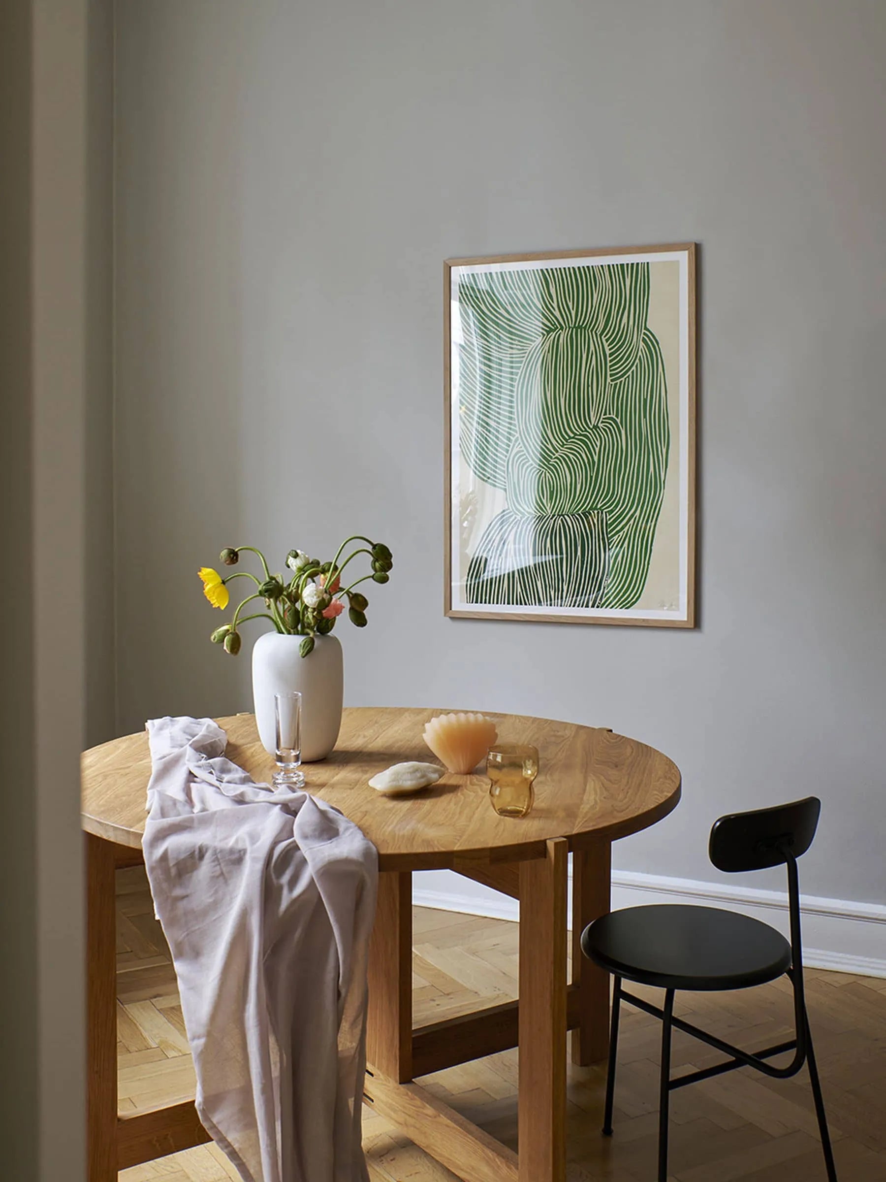 A round wooden table with a vase, two glasses, and a draped cloth sits by a black chair. On the light gray wall hangs Green Ocean (30x40) by Rebecca Hein from The Poster Club.