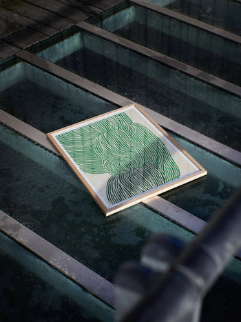 Rebecca Heins Green Ocean (30x40) from The Poster Club features green curved lines, framed and set above water on glass, with reflections and metal railings enhancing this abstract art print.