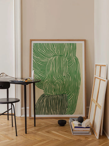 The Poster Clubs Rebecca Hein - Green Ocean (30x40) abstract artwork with green and white lines leans against a beige wall on a wooden floor, beside blank frames, books, and a black table with a chair.
