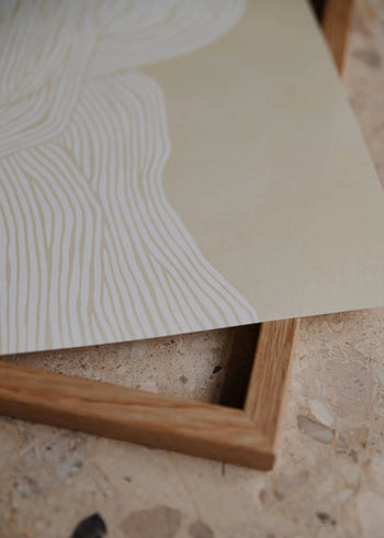 A close-up of The Poster Clubs Rebecca Hein - The Line No. 7 (30x40) art print, featuring white wavy lines on light beige, partially resting on a wooden frame atop a textured stone surface.
