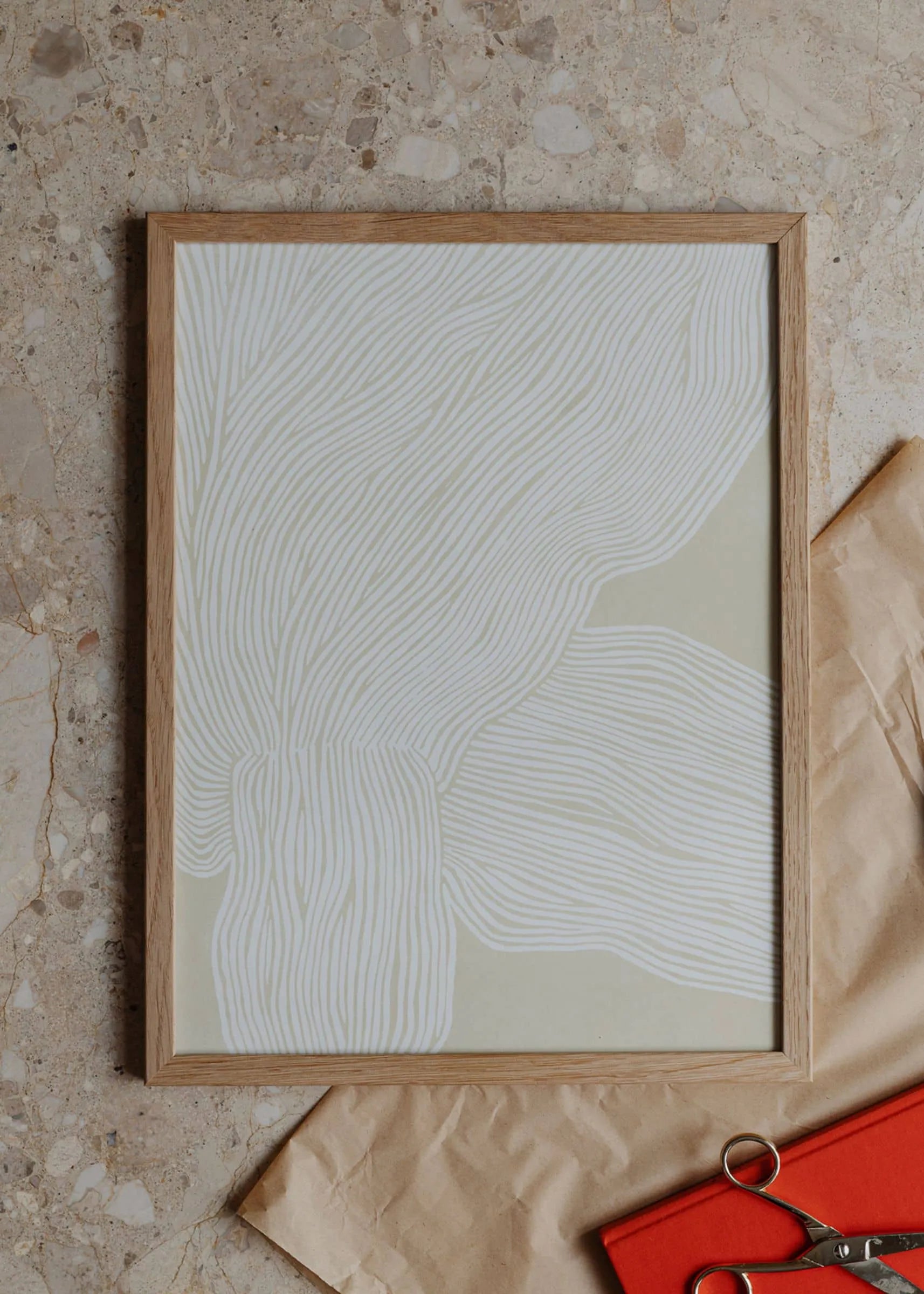 The Poster Clubs Rebecca Hein - The Line No. 9 (30x40) framed art print showcases white wavy lines on beige, styled on a speckled stone surface with brown wrapping paper, an orange notebook, and metal scissors.