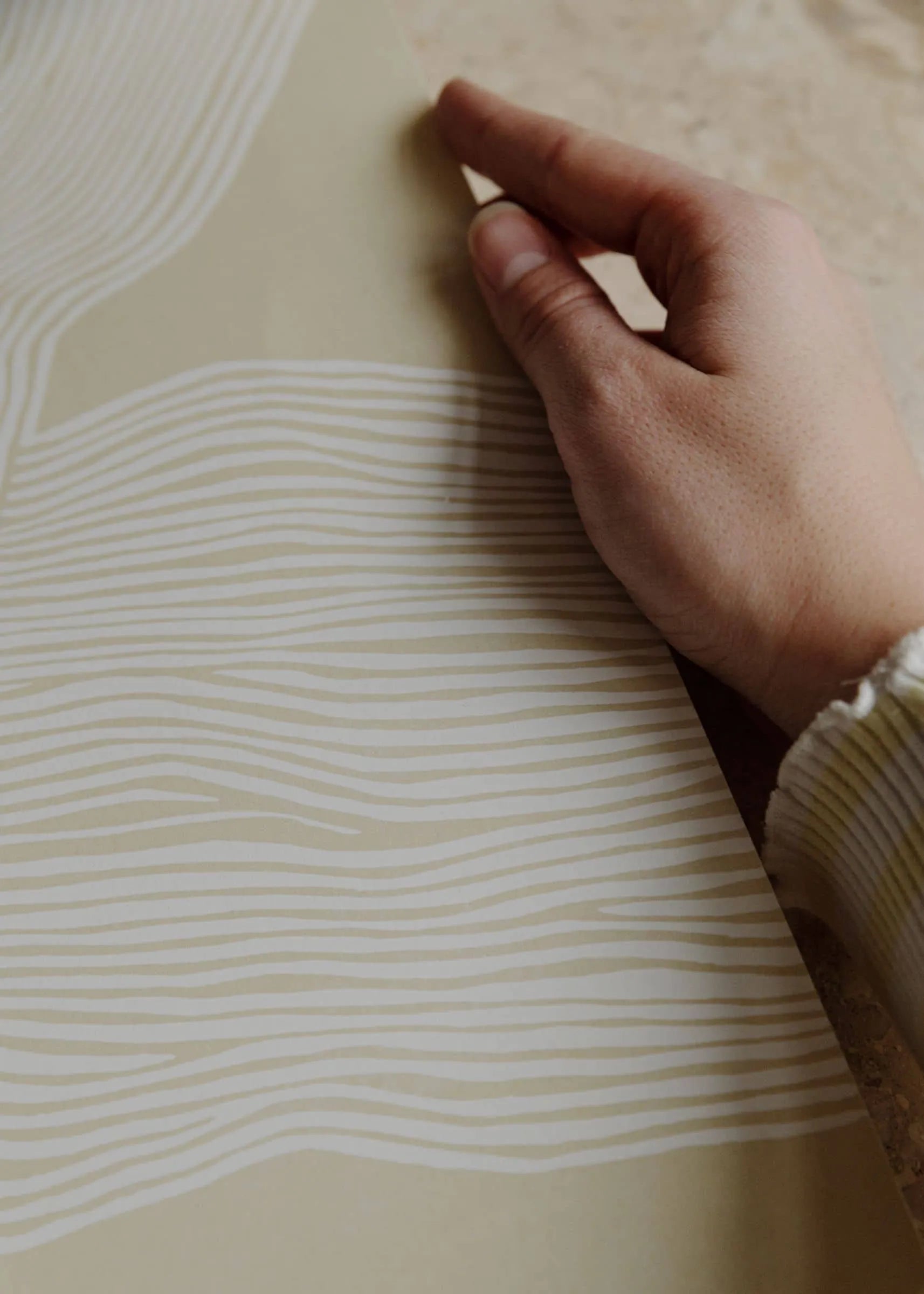 A hand holds The Poster Club’s Rebecca Hein - The Line No. 9 (30x40), a beige art print featuring wavy white lines that evoke Nordic design, reminiscent of Rebecca Hein’s style. The person wears a light, long-sleeved top.
