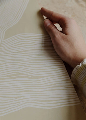 A hand holds The Poster Club’s Rebecca Hein - The Line No. 9 (30x40), a beige art print featuring wavy white lines that evoke Nordic design, reminiscent of Rebecca Hein’s style. The person wears a light, long-sleeved top.
