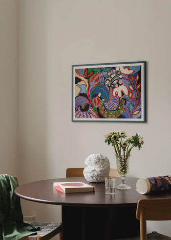 A round dark wood table with a vase of white flowers, a textured white sculpture, a pink book, and The Poster Club Rini Spiel - Felicity (50x70) giclée print sits under a colorful abstract painting. A green jacket is draped over a chair.