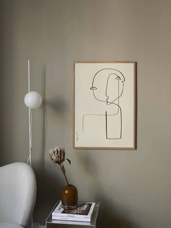 Minimalist room with a neutral wall, The Poster Club Anastasia Krasilnikova - Jaguar 01 (50x70) print in a wooden frame, white floor lamp, white chair, and clear side table with a brown vase of dried flowers and stacked books.