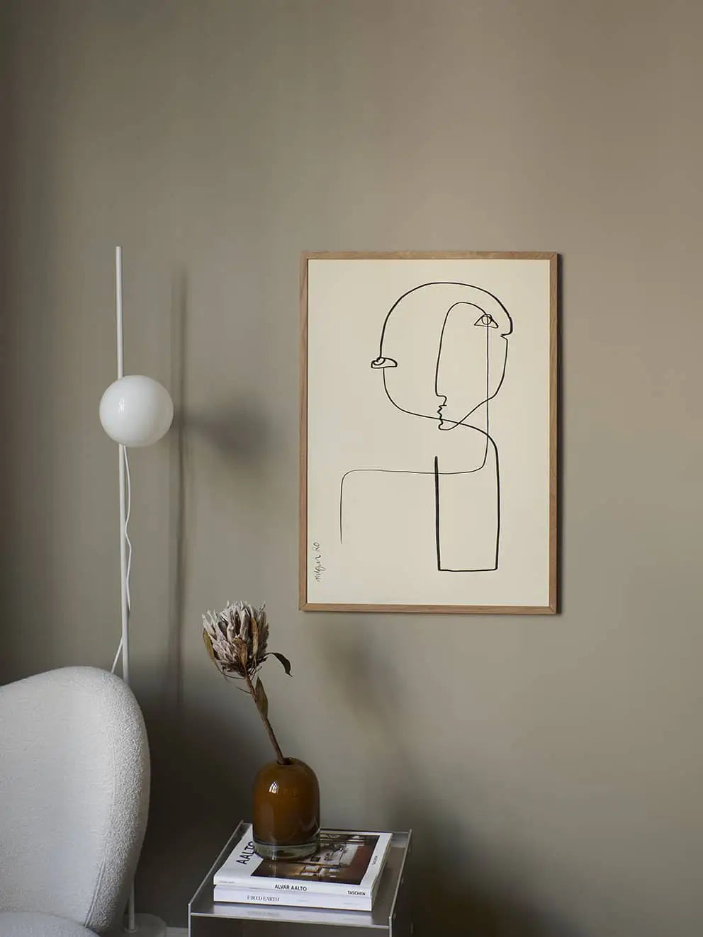 A minimalist room with a beige wall, featuring The Poster Club Anna Mörner - Statue (50x70) fine art print in a frame, a white modern lamp and chair, a brown vase with dried flowers, and books stacked on a clear side table.
