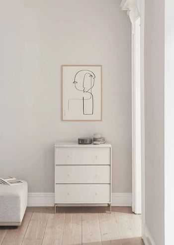 A minimalist room with a light wood floor features a white three-drawer dresser topped with stacked bowls, and The Poster Club Robin Ahlgren - Sculpture No. 02 (50x70) art print by The Poster Club framed above it, as light pours in from a large window on the right.