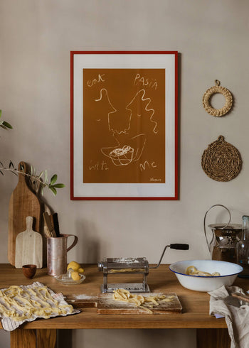 A rustic kitchen features a wooden table with a pasta maker, fresh pasta, eggs, and flour. On the wall hangs The Poster Club Ruby Hughes - Eat Pasta (30x40) art print. Woven decor and cutting boards add charm.