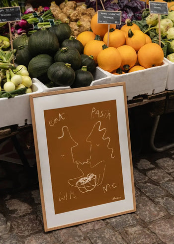 The Poster Club Ruby Hughes - Eat Pasta (50x70) print, featuring abstract white line art and the phrase eat pasta with me, is framed and rests against a vegetable stand with zucchinis, yellow melons, and other market produce.