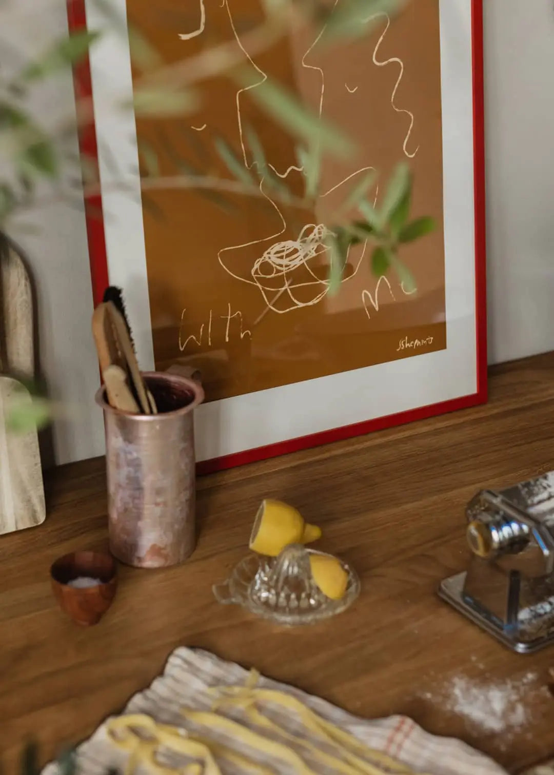 On the kitchen counter: a citrus juicer with lemon halves, a pasta machine, utensils in a metal cup, and The Poster Club Ruby Hughes - Eat Pasta (50x70) fine art giclée print framed in red in the background.