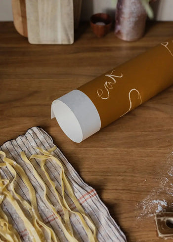 Fresh pasta strips dry on a striped cloth next to a brown rolling pin with white handles, as seen in Ruby Hughes Eat Pasta (50x70) fine art print from The Poster Club.