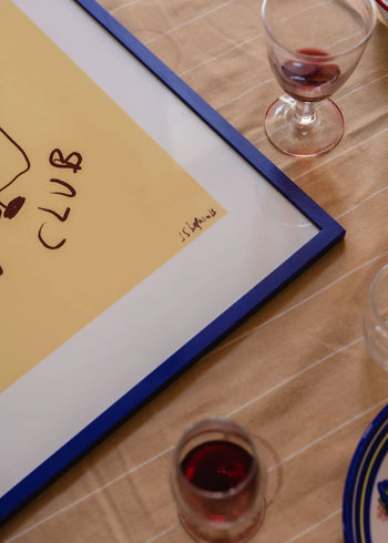 The Poster Club’s Ruby Hughes - Wine Club (30x40) giclée print, featuring a yellow background and handwritten text, rests on a beige striped tablecloth surrounded by glasses of red wine and a clear jar.