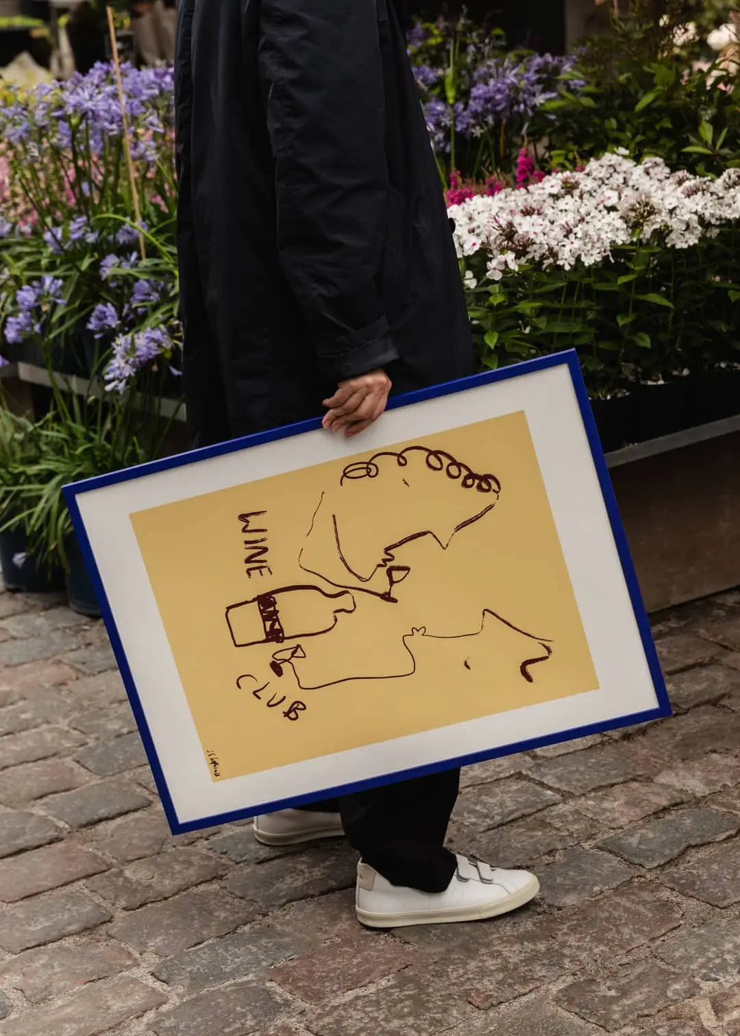 A person in white sneakers holds The Poster Club Ruby Hughes - Wine Club (50x70) print, featuring line art faces and a WINE CLUB bottle on yellow, standing on a cobblestone street near vibrant potted flowers.