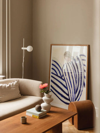 A modern living room with neutral tones features a beige sofa, wooden coffee table, and The Poster Club’s Sofia Lind - Blue Stripe At Concorde (30x40) print from the Paris Metro Collection. A distinctive white lamp stands nearby.