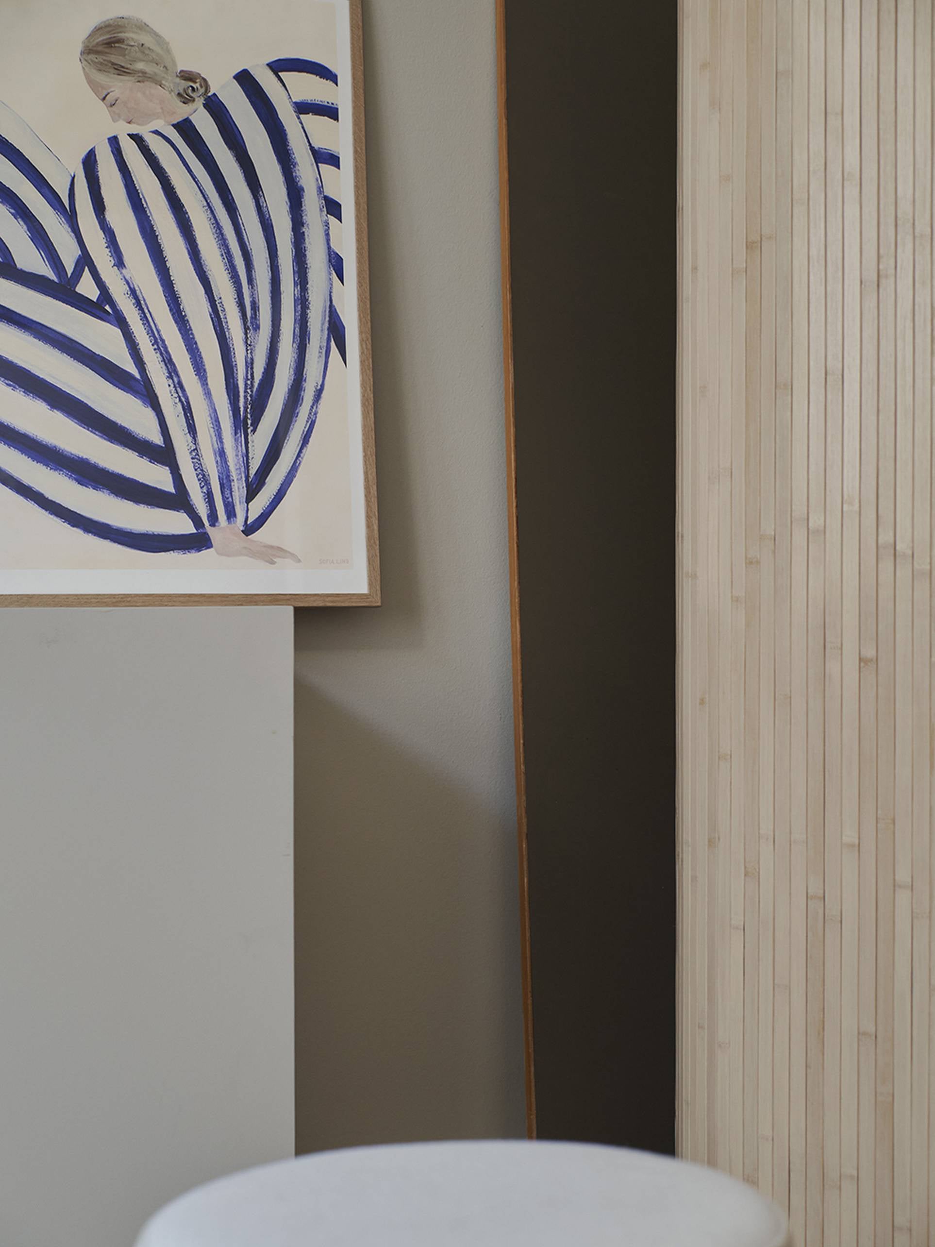 The Poster Club Sofia Lind - Blue Stripe At Concorde (50x70) Giclée print features a blonde figure in a white garment with bold blue stripes, displayed on a neutral wall next to wood paneling and a white round surface.