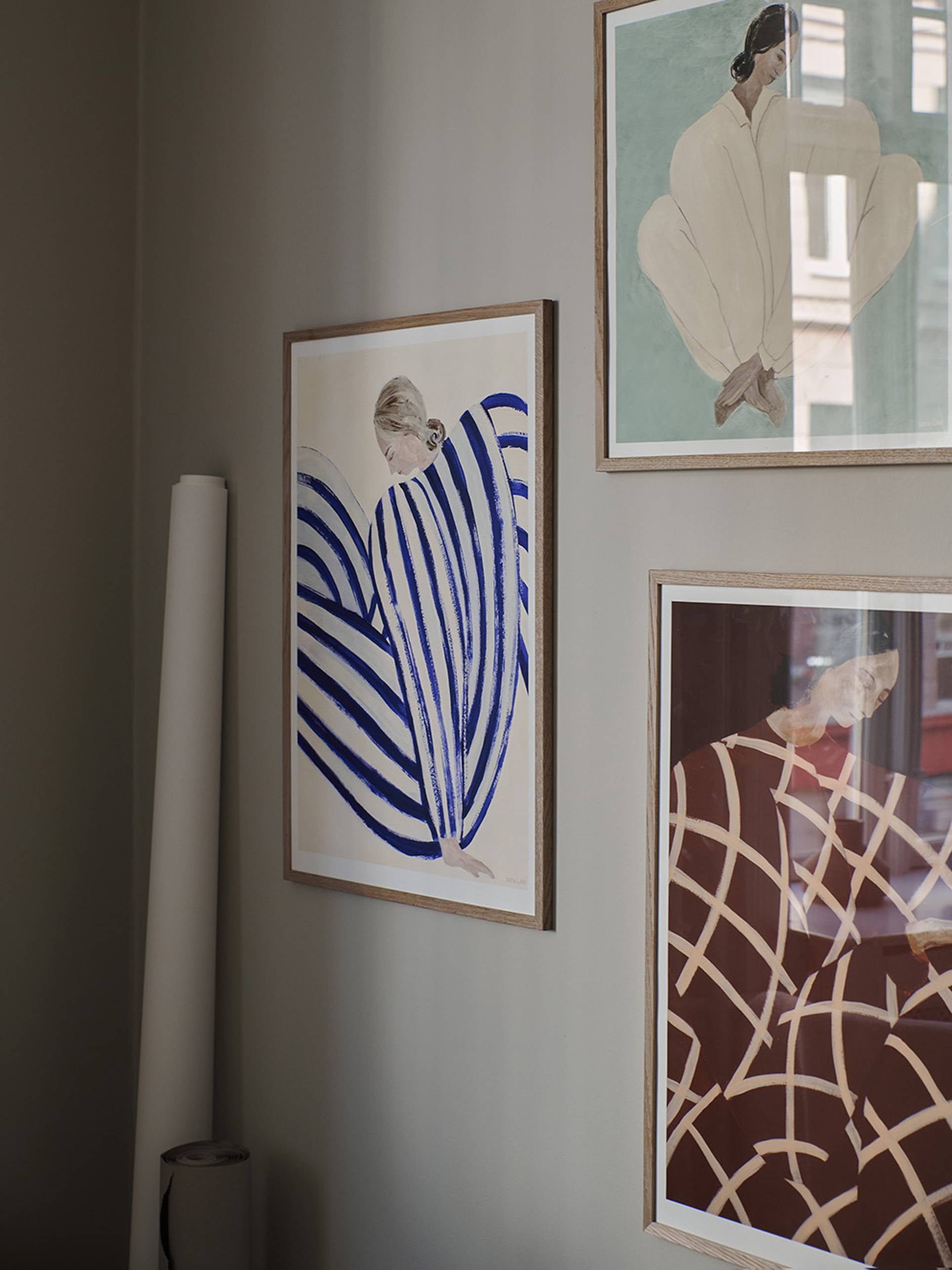 The Poster Clubs Sofia Lind - Blue Stripe At Concorde (50x70) Giclée print hangs on a light gray wall, showing a partially obscured figure among bold blue, beige, and brown shapes; a rolled paper tube rests below.